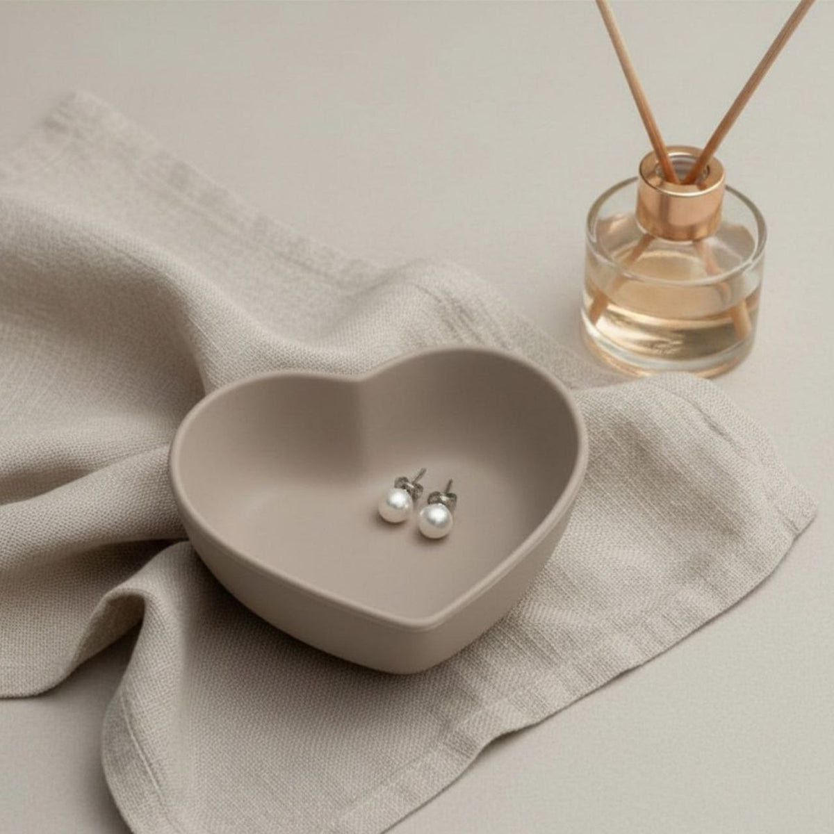 Heart trinket tray in taupe finish on a beige background and pearl earrings and a reed diffuser