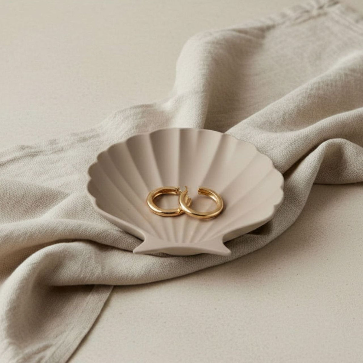 Taupe Clam Shell-shaped jewelry dish with gold ear rings on a beige fabric background