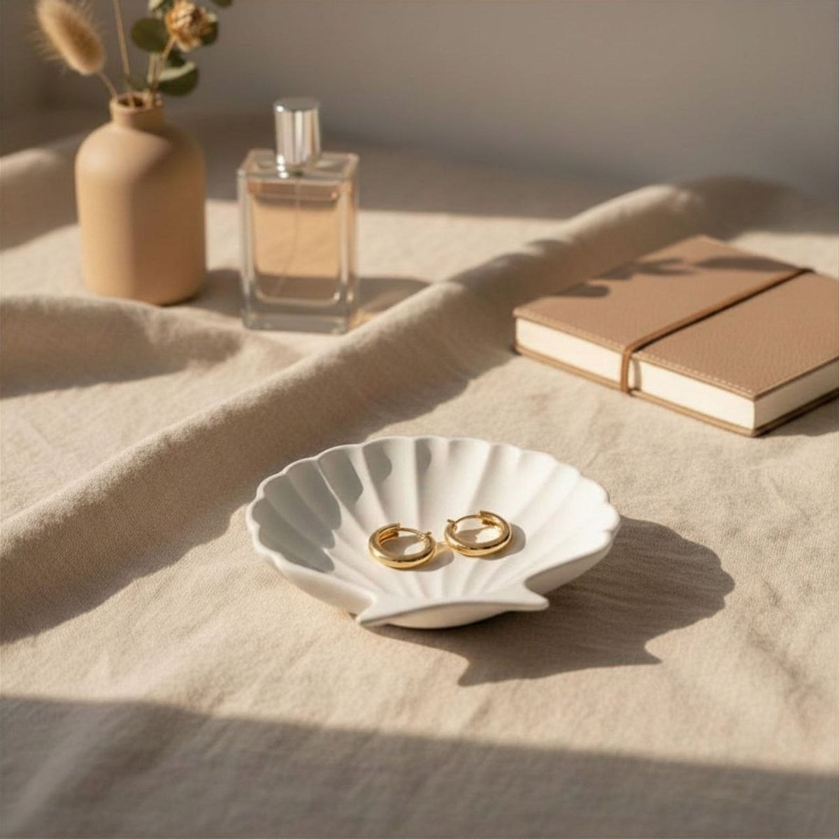 White clam shell-shaped jewelry dish with gold ear rings on a beige surface with a vase, perfume bottle, and books in the background.