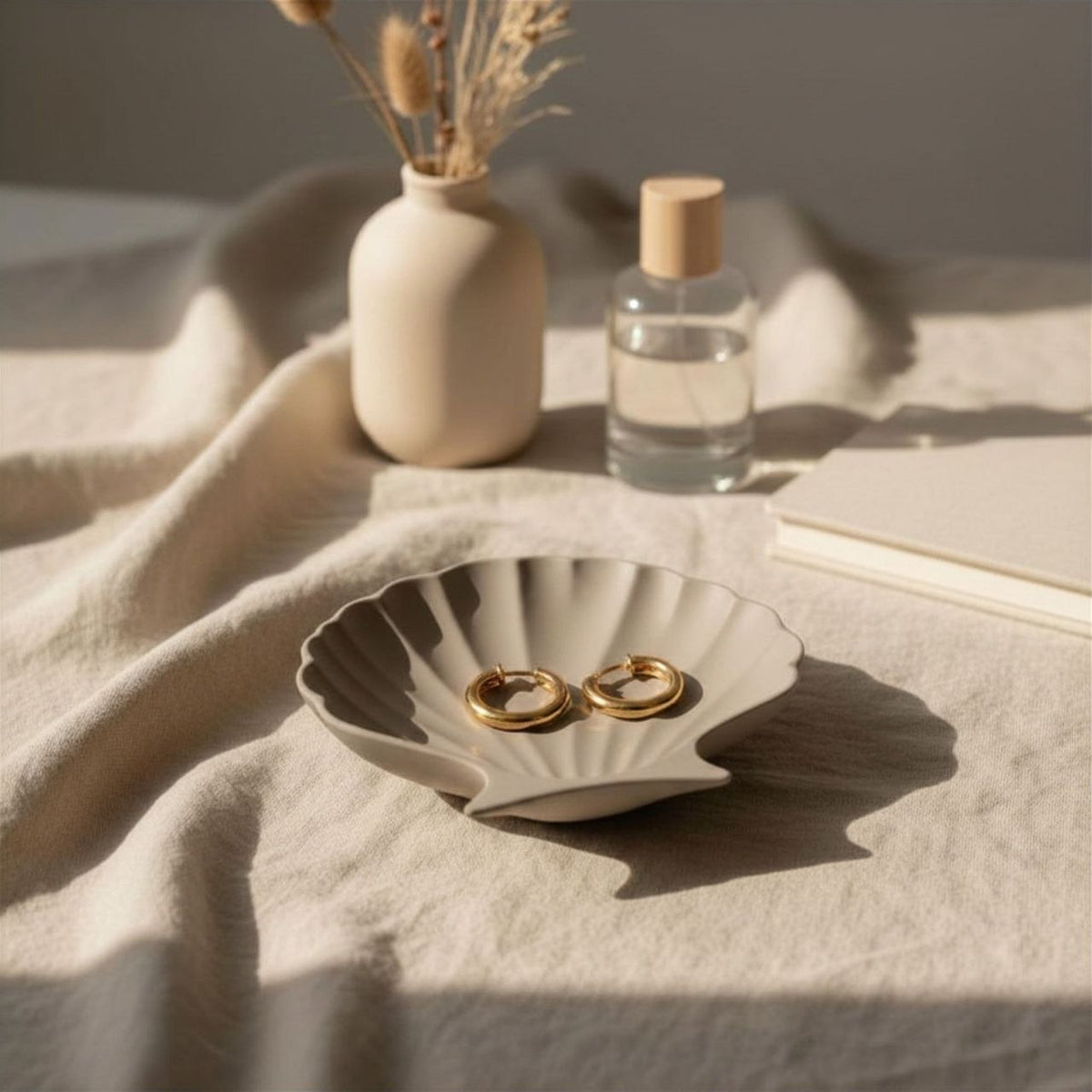 Taupe Clam Shell Jewelry dish with gold ear rings, small vase, and bottle on a textured surface