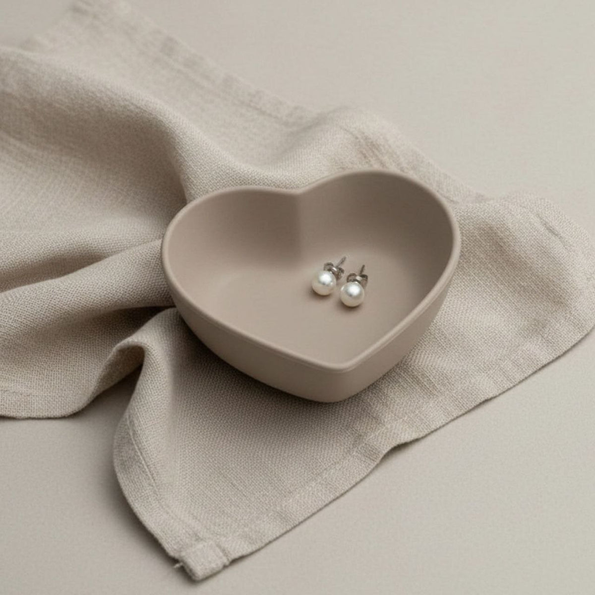Heart trinket tray in taupe finish on a beige background and pearl earrings.