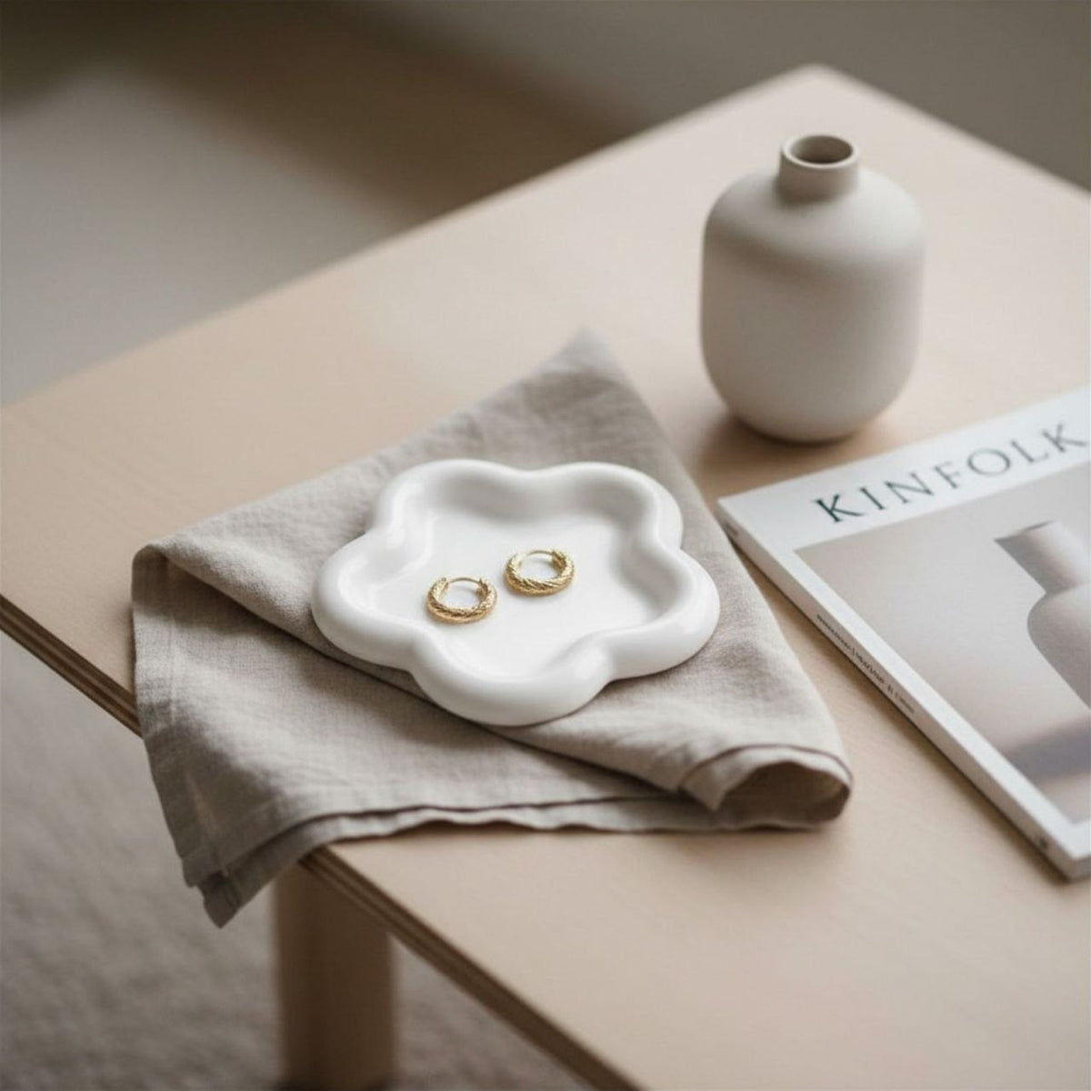 Natural finish flower tray with gold earrings on a beige cloth, next to a white vase and a magazine on a wooden surface.