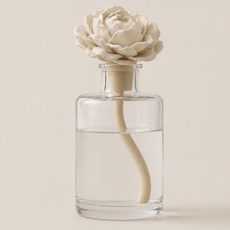 Clear glass diffuser bottle with a white floral peony topper on a light beige background