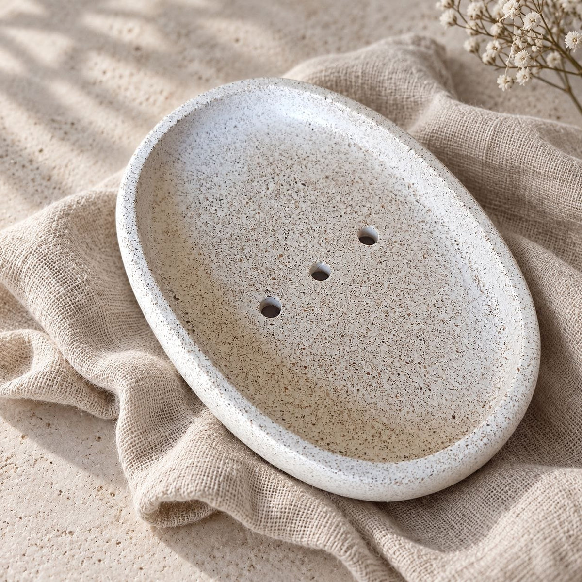 Natural oval soap dish with a speckled finish, raised rim and three drainage holes, resting on beige fabric.