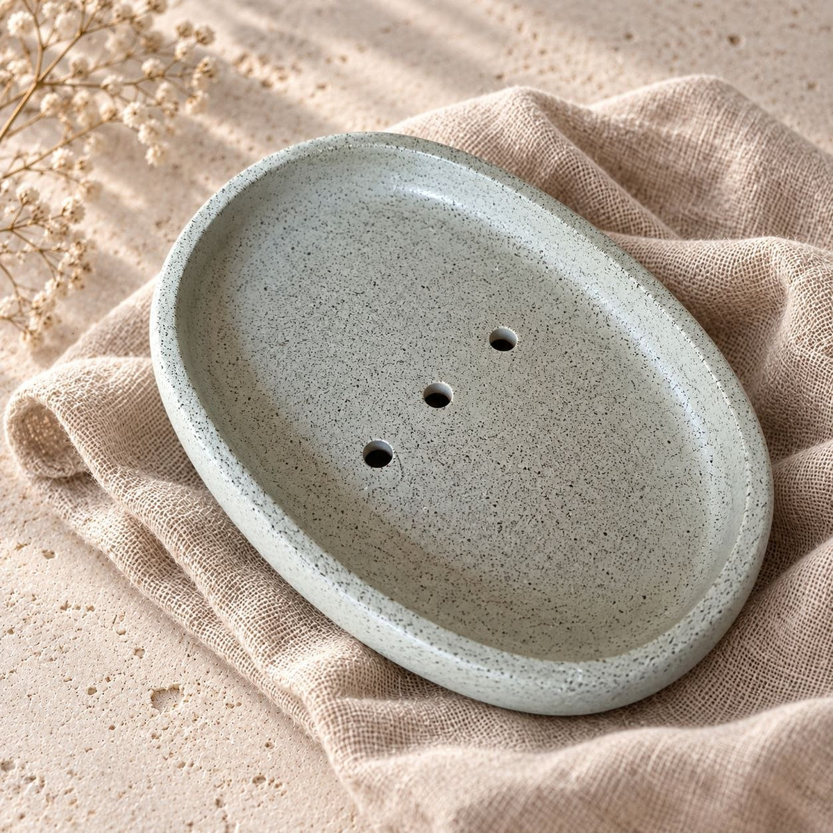 Sage Green oval soap dish with a speckled finish, raised rim and three drainage holes, resting on beige fabric.