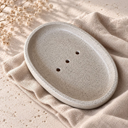 Light Taupe oval soap dish with a speckled finish, raised rim and three drainage holes, resting on beige fabric.
